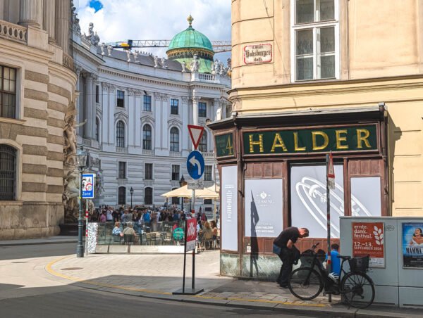 Green-domed Michaelertrakt of the Hofburg seen from the corner of Habsburgergasse in Vienna, with the gold-lettered Halder shopfront, a café terrace and crowds on Michaelerplatz