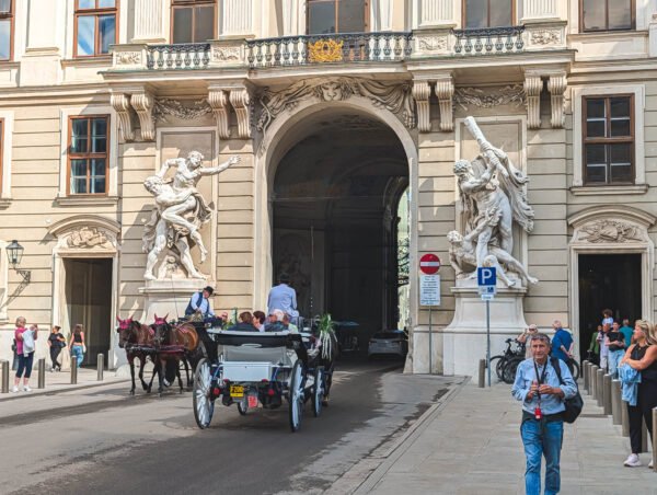 Fiaker carriage with two horses coming through an arched gateway of the Imperial Chancellery Wing (Reichskanzleitrakt) of the Hofburg, Vienna, flanked by large marble statues of Hercules in combat