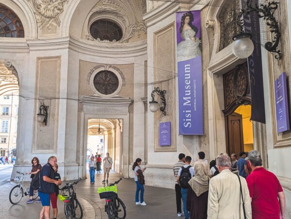 Visitors queuing at the Sisi Museum and Imperial Apartments entrance in the arched Michaelertrakt passage of the Hofburg, Vienna, with a purple banner of Empress Elisabeth and cyclists in the passage