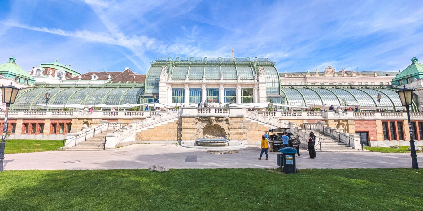 Art Nouveau glass and steel Palmenhaus seen across the lawn of the Burggarten, Vienna, with double stone stairs, a fountain niche and café terrace under a blue sky