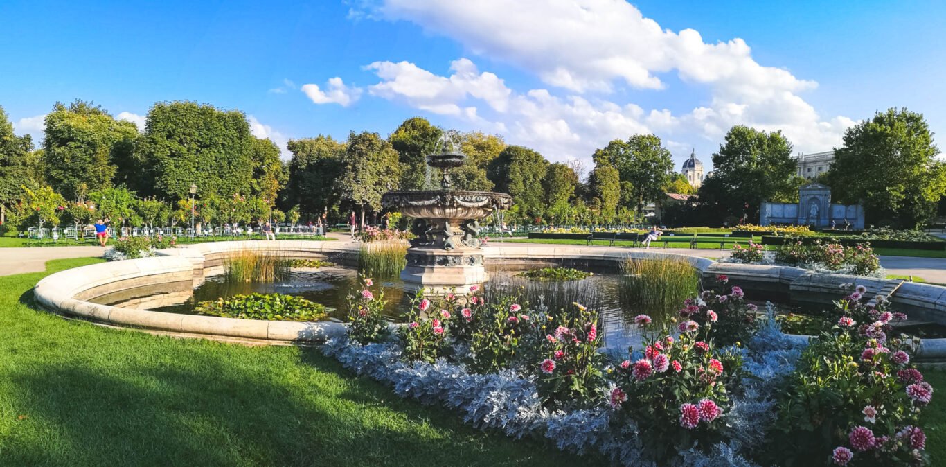 Stone fountain in a round pond surrounded by pink dahlias and silver foliage in the Volksgarten, Vienna, with trees, rose beds and the Theseus Temple under a blue sky